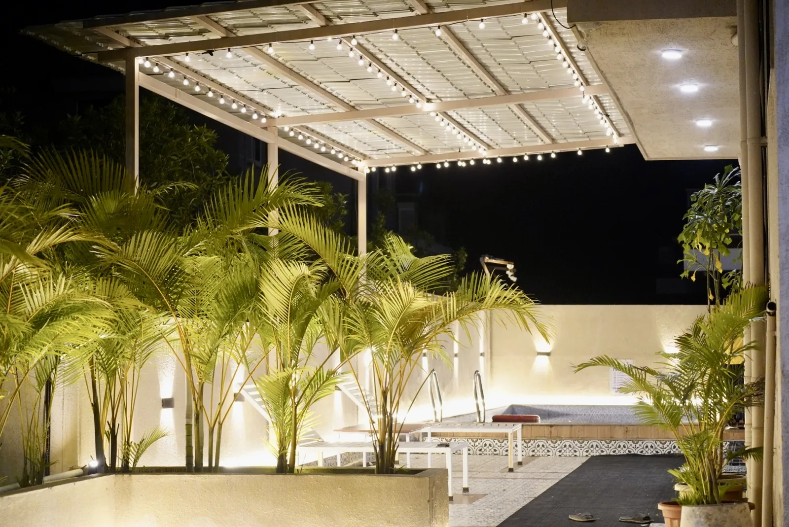 A covered deck with string lights, potted palms and white loungers beside the pool.