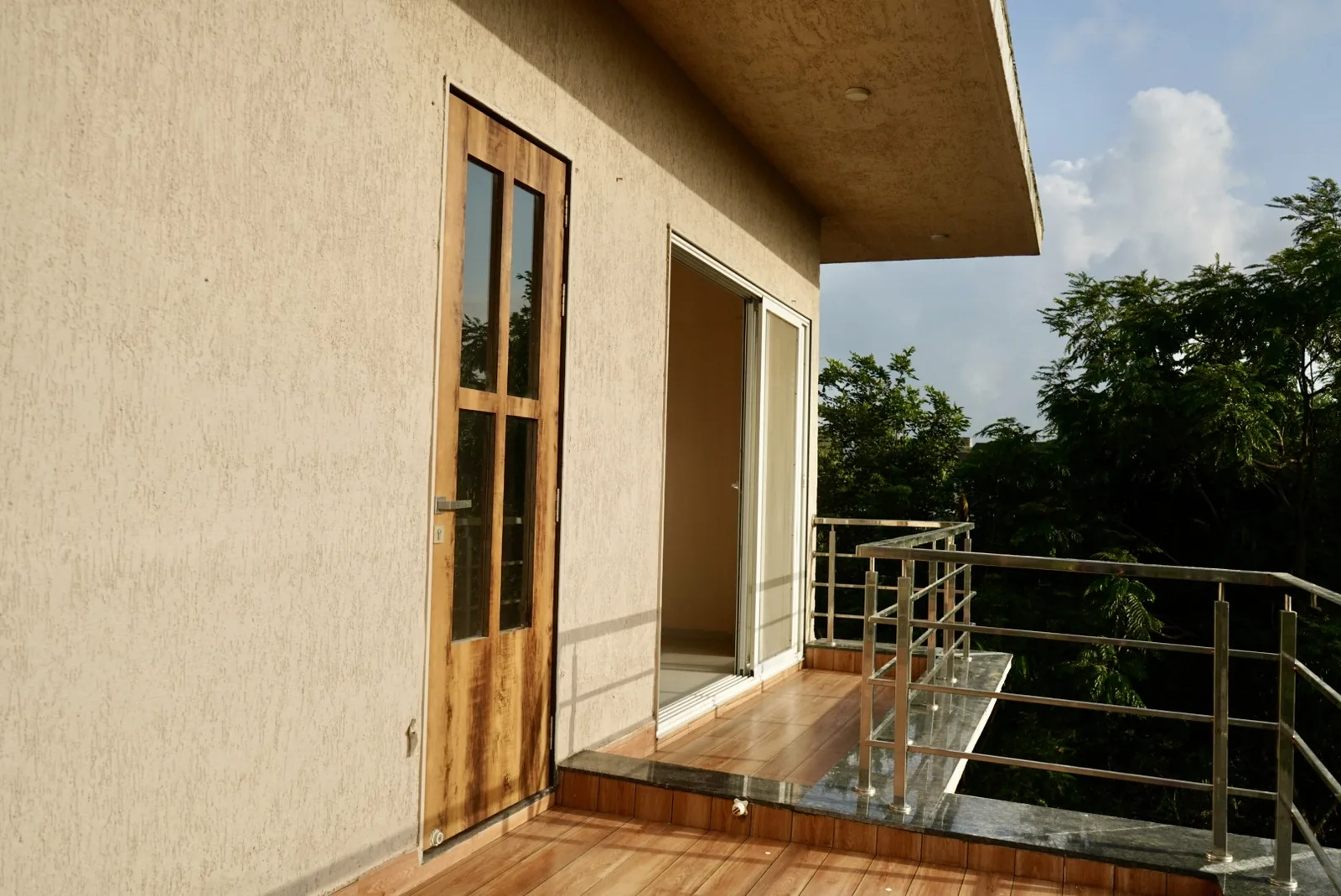 A sunlit tiled balcony with metal railings, an open sliding door and trees beyond.